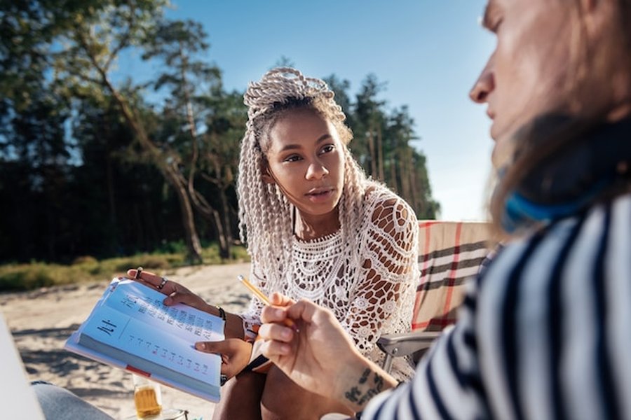 Une femme aux tresses blanches consulte un carnet de notes sur une plage pour suivre les conseils du Guide du Quiet Travel : comment planifier un voyage dans les zones de silence certifiées et les retraites de déconnexion totale.