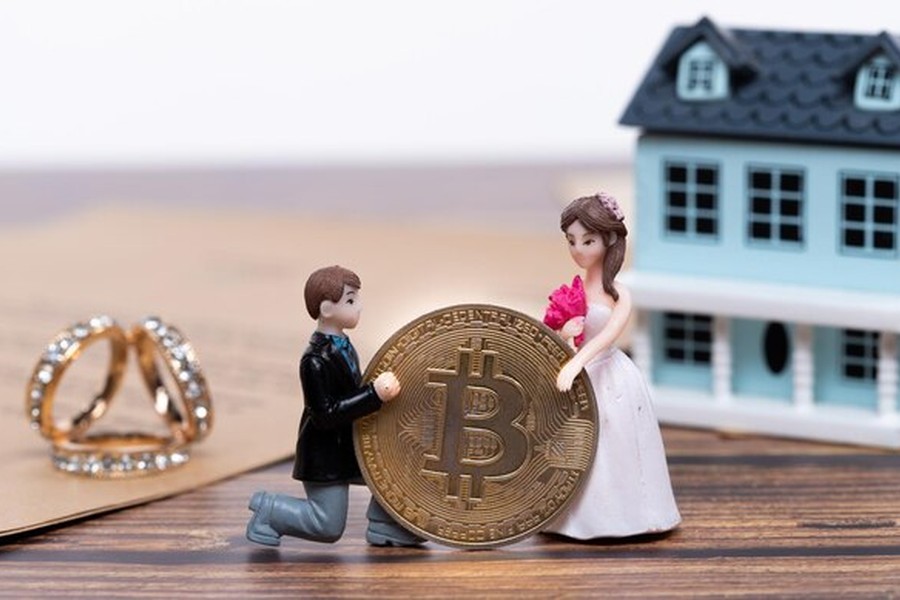 Des figurines de jeunes mariés tenant un Bitcoin devant une maison miniature et des alliances, illustrant le Mariage et actifs numériques : comment intégrer vos cryptomonnaies et NFT dans votre contrat en 2026.