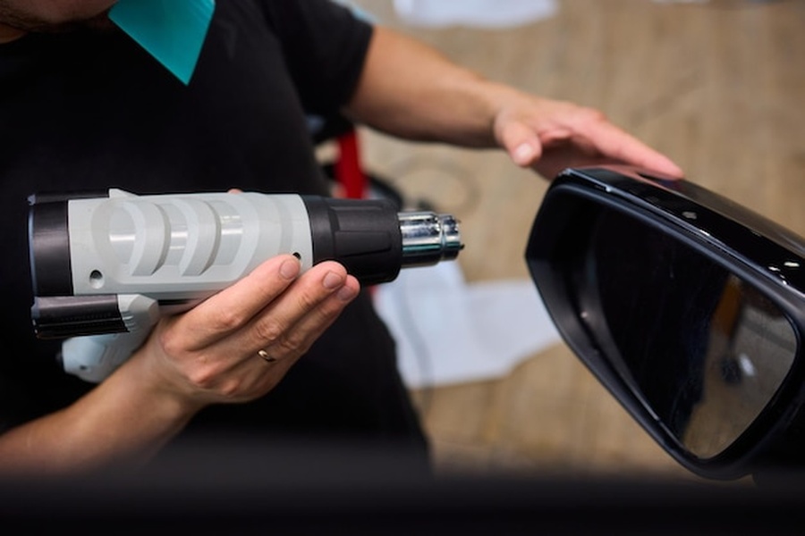 Un technicien utilise un pistolet thermique sur le rétroviseur d'une voiture électrique dans le cadre d'un Guide technique : Adapter son installation électrique pour le Vehicle-to-Home (V2H).