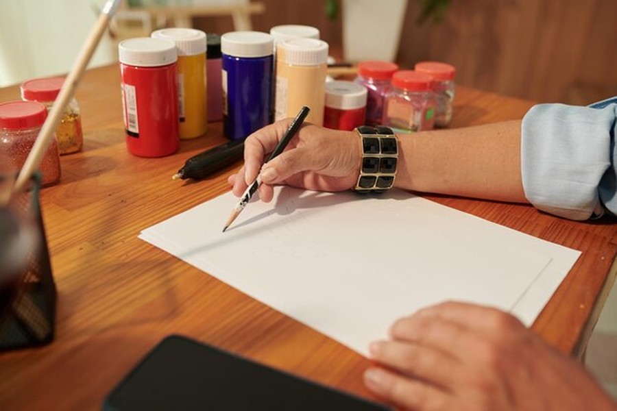Un artiste dessine au crayon sur une feuille blanche entouré de pots de peinture, illustrant comment prouver qu'une œuvre est peinte à la main pour rassurer les collectionneurs.