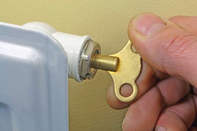 Une main utilise une petite clé de purge en laiton pour ouvrir la vis de purge située sur le côté d'un radiateur blanc.